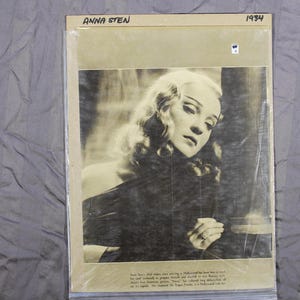 May include: Black and white photo of a woman with long, wavy hair. She is wearing a dark dress and looking down. The photo is dated 1934 and has the name "Anna Sten" printed at the top.