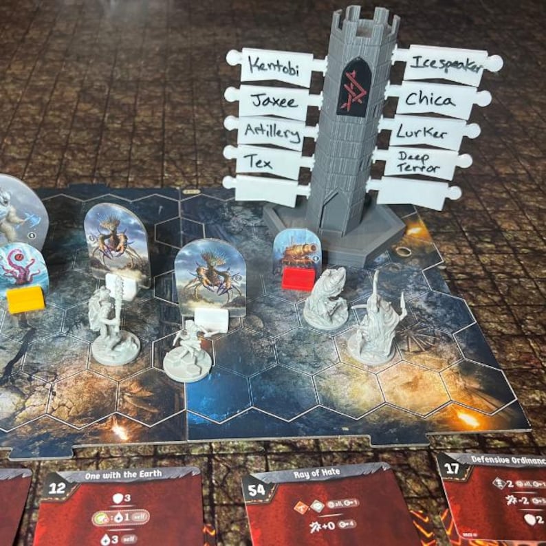 Pu&ograve; includere: Un'impostazione di gioco da tavolo con una struttura a torre grigia e carte personaggio. La torre ha etichette con nomi come "Kentobi" e "Icespeaker". Sono visibili anche pedine e carte con testo come "One with the Earth" e "Ray of Hate".