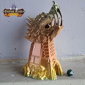 May include: A gold and bronze dice tower with a detailed, stylized monster head design. The tower has a geometric base and a multi-sided die is next to it. The tower has the text "Fates End Dice Towers".