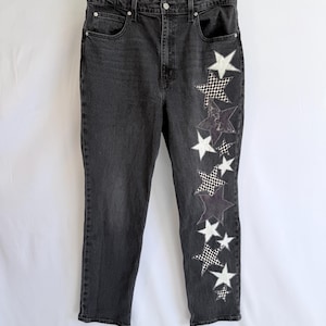 May include: Dark gray denim jeans with a high waist and straight leg. The left leg features a vertical design of white and black checkered and solid white star patches. The jeans are hanging against a white background.