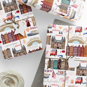 Op de afbeelding: Cadeaus verpakt met papier met een Amsterdam-thema. Het papier toont illustraties van gebouwen, bruggen, fietsen en de tekst "City of Bikes" en "Amsterdam". Cadeaus zijn vastgebonden met touw en een stervormig label. Een bol touw, een schaar en labels zijn ook zichtbaar.