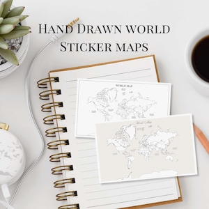 May include: A notebook with two world map sticker designs, a cup of coffee, and a pencil. The text "HAND DRAWN WORLD STICKER MAPS" is displayed above the notebook. A succulent plant and headphones are also visible.