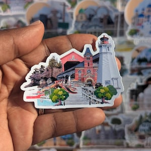 Könnte beinhalten: Ein farbenfroher Aufkleber mit einer Collage von Wahrzeichen einer Küstenstadt. Der Aufkleber zeigt einen roten Leuchtturm, ein rotes Gebäude mit weißen Verzierungen, eine Brücke, einen Hummer und grüne Bäume. Der Aufkleber ist auf weißem Papier gedruckt und hat eine glänzende Oberfläche.