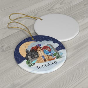 May include: Two round ceramic ornaments with gold hanging strings. One is plain white, while the other features a colorful illustration of an Icelandic landscape with a church, red house, mountains, and the text ICELAND printed in black.