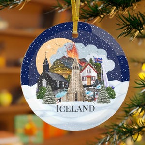 Colourful tempered glass Iceland ornament with a blue background and white snow. The baubles feature a colourful illustration of popular Icelandic Landmarks like the Reykjavik church, waterfall, volcano, black sand beach, elephant rock, puffins and more! The text "ICELAND" is printed on under the design.