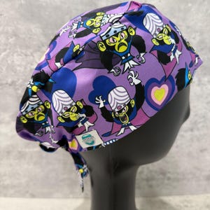 Op de afbeelding: Een paarse scrubcap met een herhalend patroon van een stripfiguur met een witte kop, gele ogen en een zwarte cape. De cap heeft een strik aan de achterkant en wordt getoond op een zwarte mannequinhoofd.