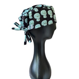 May include: A black surgical scrub cap featuring a repeating pattern of light blue cartoon faces. The cap is tied at the back with matching fabric ties. The cap is displayed on a black mannequin head against a white background.