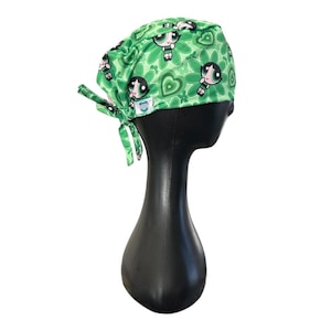 May include: Green scrub cap featuring a repeating pattern of cartoon characters and floral designs. The cap is tied at the back and displayed on a black mannequin head. The fabric has a light green background with darker green accents.