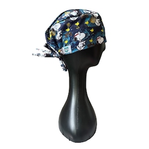 May include: A navy blue surgical scrub cap featuring a Snoopy cartoon print. The cap has a tie-back design and is displayed on a black mannequin head. The Snoopy print includes images of Snoopy, stars, and waves.