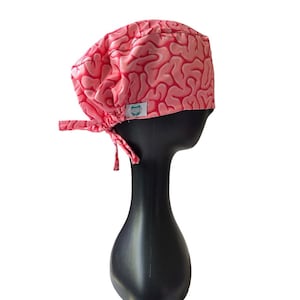 May include: A pink scrub cap featuring a brain pattern in a darker pink. The cap is tied at the back with matching fabric ties. A small white tag with a logo is sewn on the side. The cap is displayed on a black mannequin head.