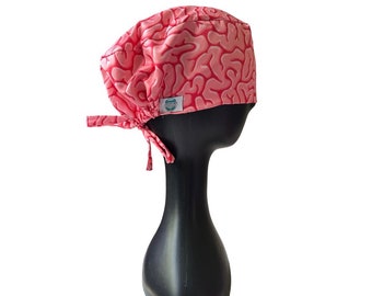 Könnte beinhalten: Eine rosa OP-Haube mit einem Gehirnmuster in dunklerem Rosa. Die Haube wird hinten mit passenden Stoffbändern gebunden. Ein kleines weißes Etikett mit einem Logo ist an der Seite angenäht. Die Haube wird auf einem schwarzen Schaufensterkopf präsentiert.