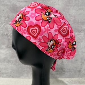 Könnte beinhalten: Eine rosa OP-Haube mit einem sich wiederholenden Muster aus Zeichentrickfiguren, Blumen und Herzen. Die Haube wird hinten gebunden und soll das Haar bedecken. Die Farbpalette umfasst Rosa-, Rot- und Orangetöne.