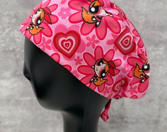 May include: A pink scrub cap featuring a repeating pattern of cartoon characters, flowers, and hearts. The cap is tied at the back and is designed to cover the hair. The color palette includes shades of pink, red, and orange.