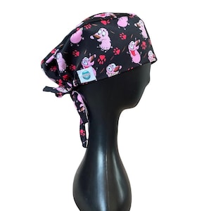 Puede incluir: Gorro quirúrgico negro con un estampado repetido de personajes de perros de dibujos animados rosas y huellas de patas rojas. El gorro está atado en la parte posterior y tiene una pequeña etiqueta blanca con un logotipo azul y blanco.