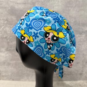Peut inclure: Un bonnet de chirurgien bleu avec un motif répété de personnages de dessins animés, de fleurs et de cœurs. Le bonnet est noué dans le dos et est fait d'un tissu léger. Le motif comprend des nuances de bleu, de jaune et de rose.