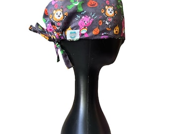 Pode incluir: Uma touca cirúrgica cinza escuro com um padrão colorido com tema de Halloween. O padrão inclui ursos de desenho animado, abóboras e raios. A touca é amarrada nas costas com uma gravata de tecido combinando.