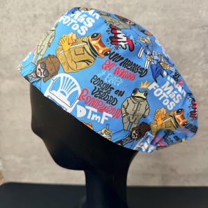 May include: A light blue scrub cap with a repeating pattern of cartoon characters, frogs, and text. The text includes the words "Más Fotos" and other phrases. The cap is designed to be worn in a medical or professional setting.