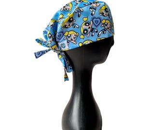 May include: A light blue scrub cap featuring a repeating pattern of cartoon characters in yellow, black, and white. The cap has a tie closure and is displayed on a black mannequin head. The design includes hearts and the cartoon characters.