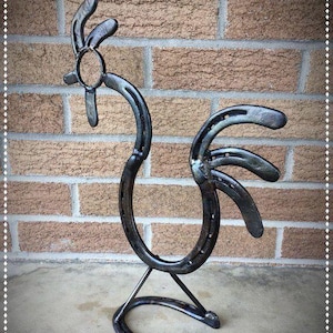 May include: A metal rooster sculpture crafted from horseshoes. The sculpture features a curved body, a circular head, and a tail made of multiple horseshoes. The rooster stands on a horseshoe base, with a brick wall in the background.