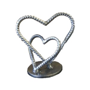 May include: A silver metal sculpture of two intertwined hearts. The hearts are made of twisted metal and are connected at the base. The sculpture is on a round base.