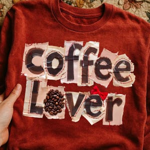 Könnte beinhalten: Rostfarbenes Sweatshirt mit den Worten "Coffee Lover" im Patchwork-Stil appliziert. Das Wort "Coffee" ist in großen Buchstaben geschrieben, wobei Kaffeebohnen das "O" im Wort "Lover" ersetzen. Eine kleine rote Schleife ist angebracht.