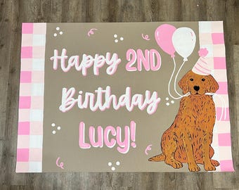 May include: A birthday sign with the text "Happy 2nd Birthday Lucy!" in white and pink. A cartoon golden retriever wearing a party hat is holding two balloons. The sign has pink and white checkered borders.