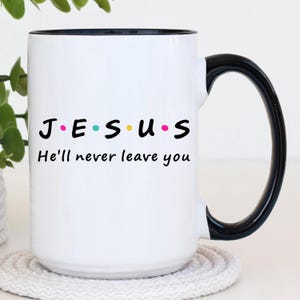May include: White ceramic mug with a black handle and rim. The mug features the word "JESUS" in black letters, with each letter in a different color. Below the word is the phrase "He'll never leave you."