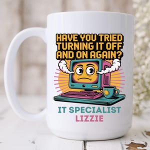 Könnte beinhalten: Weiße Keramik-Tasse mit einem Cartoon-Computer-Design und dem Text "HAVE YOU TRIED TURNING IT OFF AND ON AGAIN?" und "IT SPECIALIST LIZZIE". Das Design umfasst einen Computer, eine Tastatur und eine Kaffeetasse.