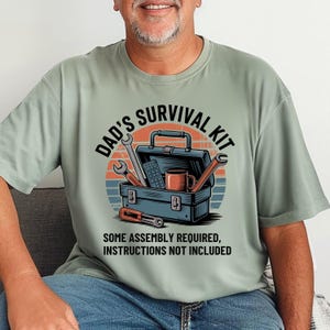 May include: Sage green t-shirt featuring a graphic of a toolbox with tools, a mug, and a remote control. The text reads "DAD'S SURVIVAL KIT" above the toolbox and "SOME ASSEMBLY REQUIRED, INSTRUCTIONS NOT INCLUDED" below.
