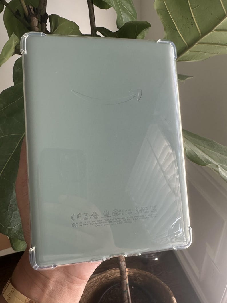 Puede incluir: Una tableta de color verde salvia dentro de una funda protectora transparente y flexible. La parte posterior del dispositivo presenta un logotipo de sonrisa de Amazon sutilmente grabado y texto reglamentario cerca del borde.