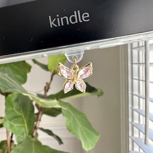 Puede incluir: Un charm de mariposa rosa y dorado cuelga de una funda de plástico transparente con la palabra "kindle" impresa.