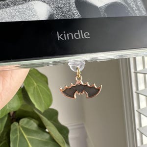 Puede incluir: Una lectora electrónica negra con la palabra "kindle" en la parte delantera. Un amuleto de murciélago dorado está unido a la lectora electrónica.