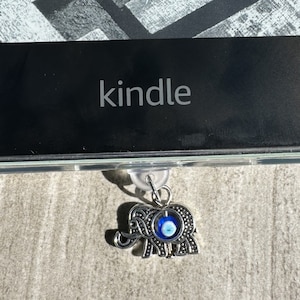 May include: A silver elephant charm with a blue evil eye charm hanging from a Kindle e-reader.