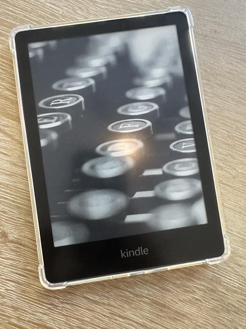 Puede incluir: Un lector electr&oacute;nico Kindle negro con una funda protectora transparente. La pantalla muestra una fotograf&iacute;a en escala de grises de alto contraste de teclas de m&aacute;quina de escribir. La palabra kindle aparece en letras blancas en el marco.