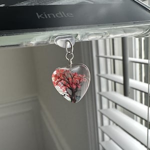 May include: A heart-shaped charm with a clear resin background and a pink flower inside. The charm is attached to a silver chain and hangs from a clear plastic hook.