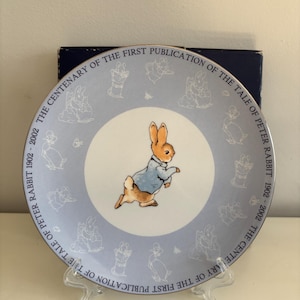 Puede incluir: Un plato de cerámica azul y blanco con una imagen de Peter Rabbit con una chaqueta azul. El plato está decorado con un borde de ilustraciones de Peter Rabbit y el texto "THE CENTENARY OF THE FIRST PUBLICATION OF THE TALE OF PETER RABBIT 1902-2002 THE CENTENARY OF THE FIRST PUBLICATION OF THE TALE OF PETER RABBIT 1902-2002".