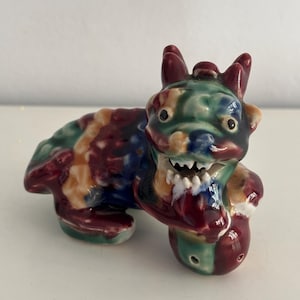 Puede incluir: Figura de cerámica colorida de una criatura mítica. La figura presenta una mezcla de colores que incluyen rojo, verde, azul y amarillo. Tiene una cara detallada con dientes afilados y pequeños cuernos.