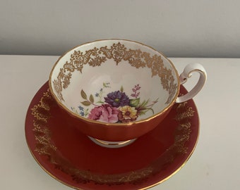 May include: A vintage teacup and saucer set. The teacup and saucer are a deep red color with gold trim. The inside of the teacup is white with a floral design in shades of pink, purple, and blue. The teacup has a curved handle.