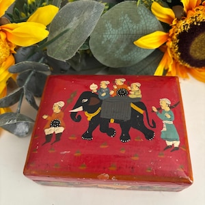 May include: A rectangular red box featuring a hand-painted scene of a black elephant carrying three figures on its back, with two additional figures walking alongside. The lid is decorated with small green triangular motifs on a red background.