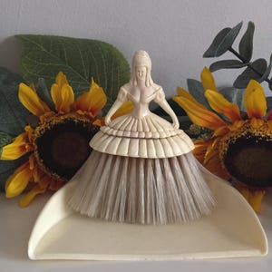 May include: A vintage dustpan and brush set. The brush is shaped like a woman in a dress, with a cream-colored figure and a tan brush. The dustpan is cream-colored. Sunflowers and greenery are in the background.