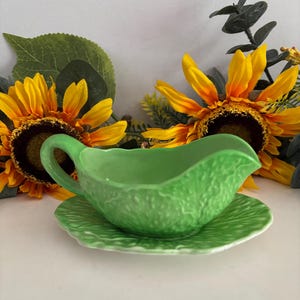 May include: A bright green ceramic gravy boat with a textured leaf-like surface and a matching saucer. The gravy boat features a curved handle and a pouring spout, designed with a detailed embossed pattern that mimics the veins of a cabbage leaf.