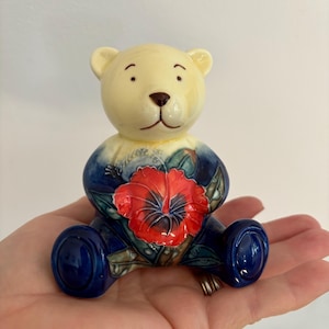 May include: A ceramic teddy bear figurine with a cream-colored head and a dark blue body. The bear has a red flower on its chest. The figurine is sitting on a hand.
