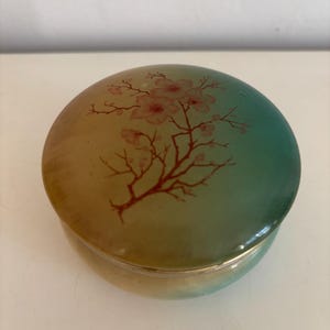 Puede incluir: Una caja de baratijas redonda con tapa, que presenta un degradado de color de oro a verde azulado. La tapa está decorada con un diseño de rama floral roja. La caja es probablemente de cerámica o vidrio.