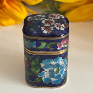 Puede incluir: Una pequeña caja rectangular de cloisonné azul con diseños florales. La caja presenta un fondo azul oscuro con detalles dorados y flores coloridas en tonos rosa, blanco y azul. La tapa y los lados están decorados con intrincados patrones florales.