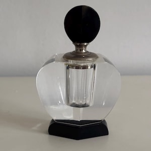 Könnte beinhalten: Ein klarer, facettierter Glasparfümflakon mit einem schwarzen, kreisförmigen Verschluss. Die Flasche hat einen silberfarbenen Metallkragen und steht auf einem schwarzen, geometrischen Sockel. Das Design der Flasche ist vintage und elegant.