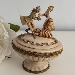 Op de afbeelding: Een sierlijke sieradendoos met deksel, met een gebeeldhouwde scène van een man en een vrouw in historische kleding. De doos is crèmekleurig met goudkleurige details en een voetstuk. De figuren zijn gedetailleerd beschilderd.