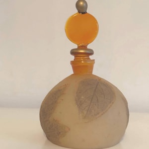 Könnte beinhalten: Ein Parfümflakon aus Milchglas mit einer kugelförmigen Basis, die mit geprägten Blattmustern versehen ist. Der Verschluss ist mit einer durchscheinenden bernsteinfarbenen Glasscheibe und einer kleinen Metallkugel gekrönt.