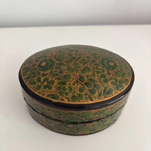 May include: An oval box with a green floral pattern on a gold base. The box has a black rim and lid. The design is intricate and the box is likely for storing small items, such as jewellery.