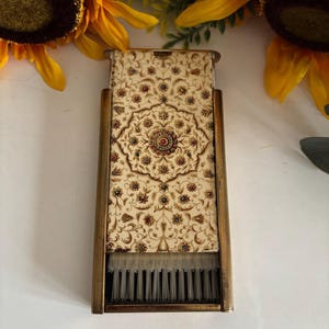 May include: A vintage vanity brush with a decorative floral pattern in cream, brown, and red. The brush has a gold-toned metal frame and a sliding cover. The bristles are white. The brush is rectangular and appears to be in good condition.