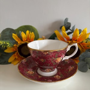Può includere: Un set di tazza e piattino da tè bordeaux con motivo floreale e bordi dorati. La tazza ha un interno bianco e un manico curvo. Il piattino è rotondo e si abbina al design della tazza. Il set è esposto con girasoli.